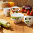 Set of ceramic bowls and cups with vegetable designs on a wooden table.