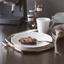 Café setting with espresso machine, cup, plate with dessert, and napkin on a reflective surface.