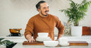 Man in a kitchen with stacks of white bowls and plates on a counter.