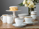 Set of white cups with gold trim on a wooden table with a cake and flowers in the background.